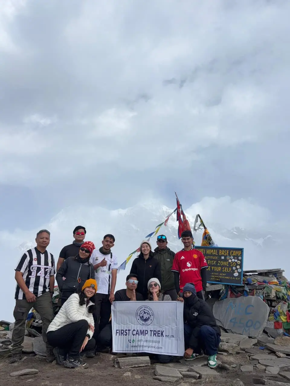 A group of trekkers posing with a First Camp Trek banner at Mardi Himal Base Camp in Nepal, next to the altitude sign post with snow-capped mountains and prayer flags in the background.
