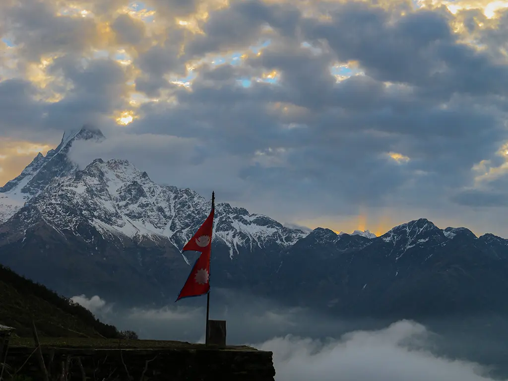 The national flag of Nepal flying on a ridge with Mount Fishtail (Machapuchare) and the Annapurna mountain range in the background under stormy sunset clouds.