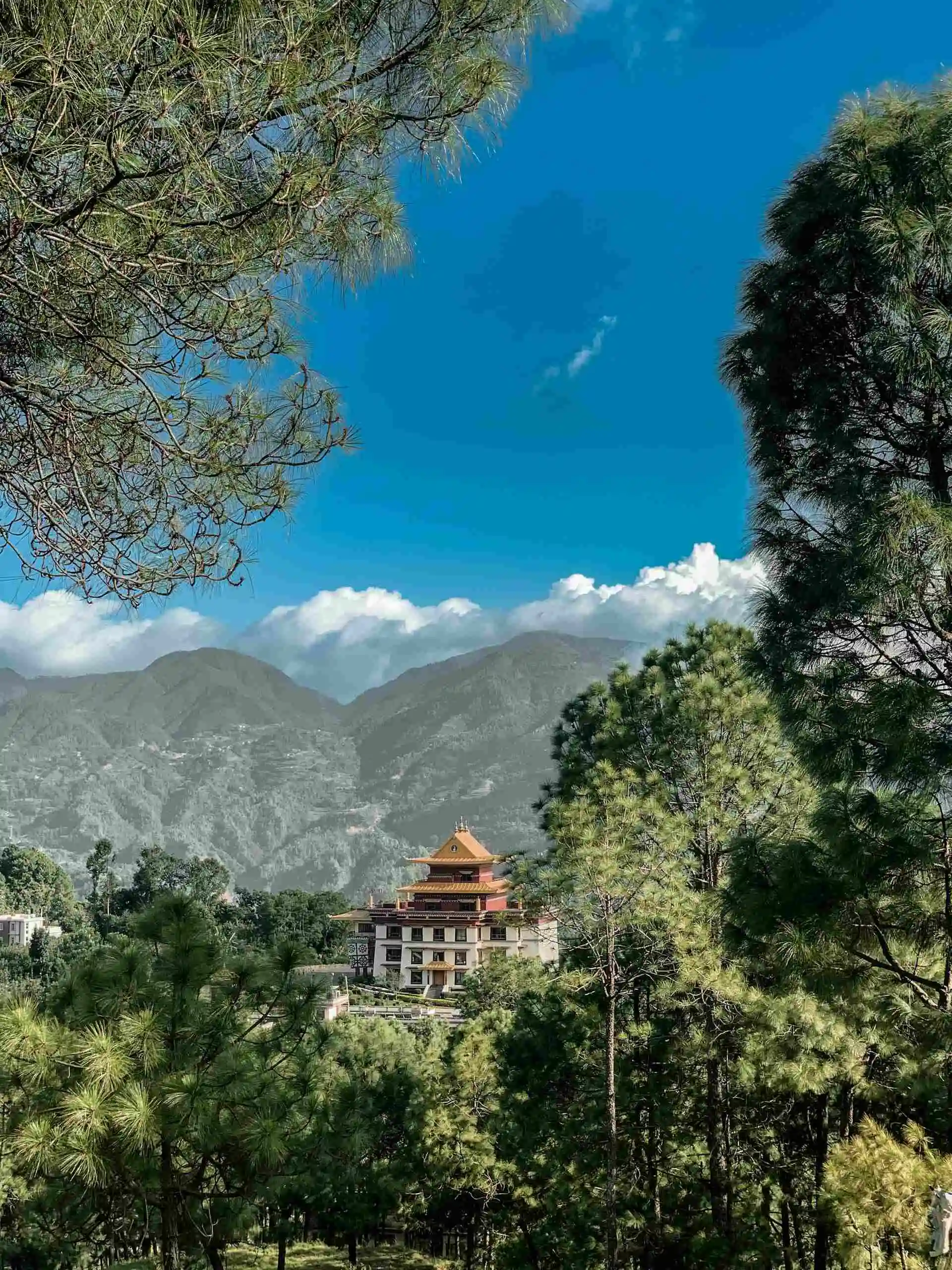 A serene view from the Champadevi hiking trail in Nepal, where a Buddhist monastery is seen nestled between trees in the foreground. Behind the monastery, a hill rises, adding to the peaceful landscape. Above, the sky is bright with a few scattered clouds, capturing the beauty of hiking in Nepal.