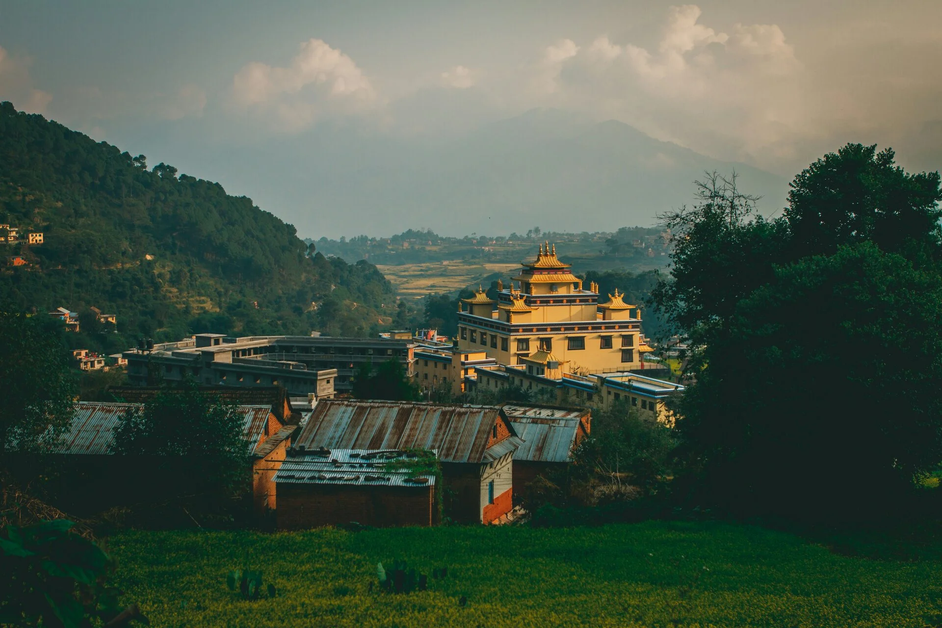A scenic view from a higher elevation captures a yellow Buddhist monastery along with a few traditional Nepali houses. Behind the monastery, small green fields stretch toward a hill covered in greenery. Some trees are scattered throughout the scene. The sky is cloudy, adding a calm atmosphere to the landscape as seen during hiking to Nepal.
