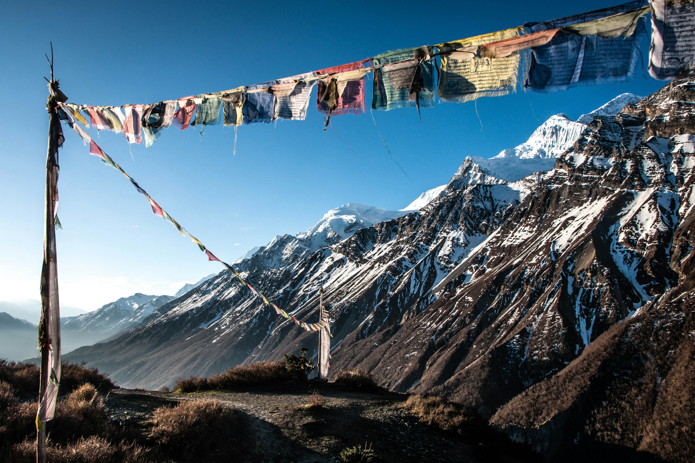 Snow-capped mountains in the background with colorful Buddhist prayer flags fluttering in the foreground, seen from Manang, Nepal, under a bright, clear sky.
