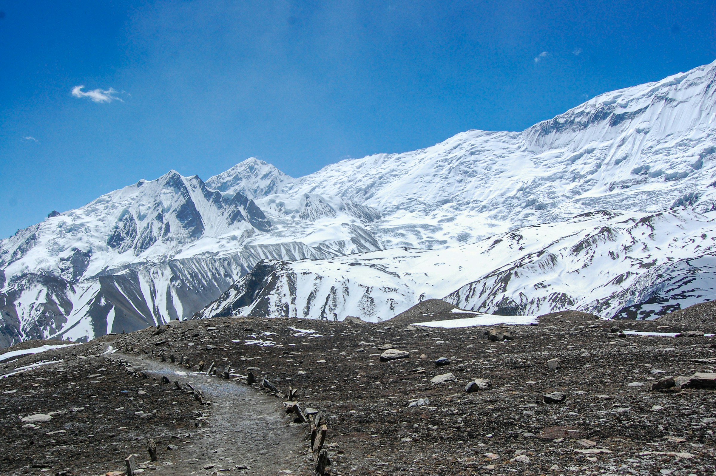 Snow-capped mountains towering in the background under a clear blue sky, overlooking a rugged offroad route to Manang, Nepal, surrounded by barren fields and rocky terrain.