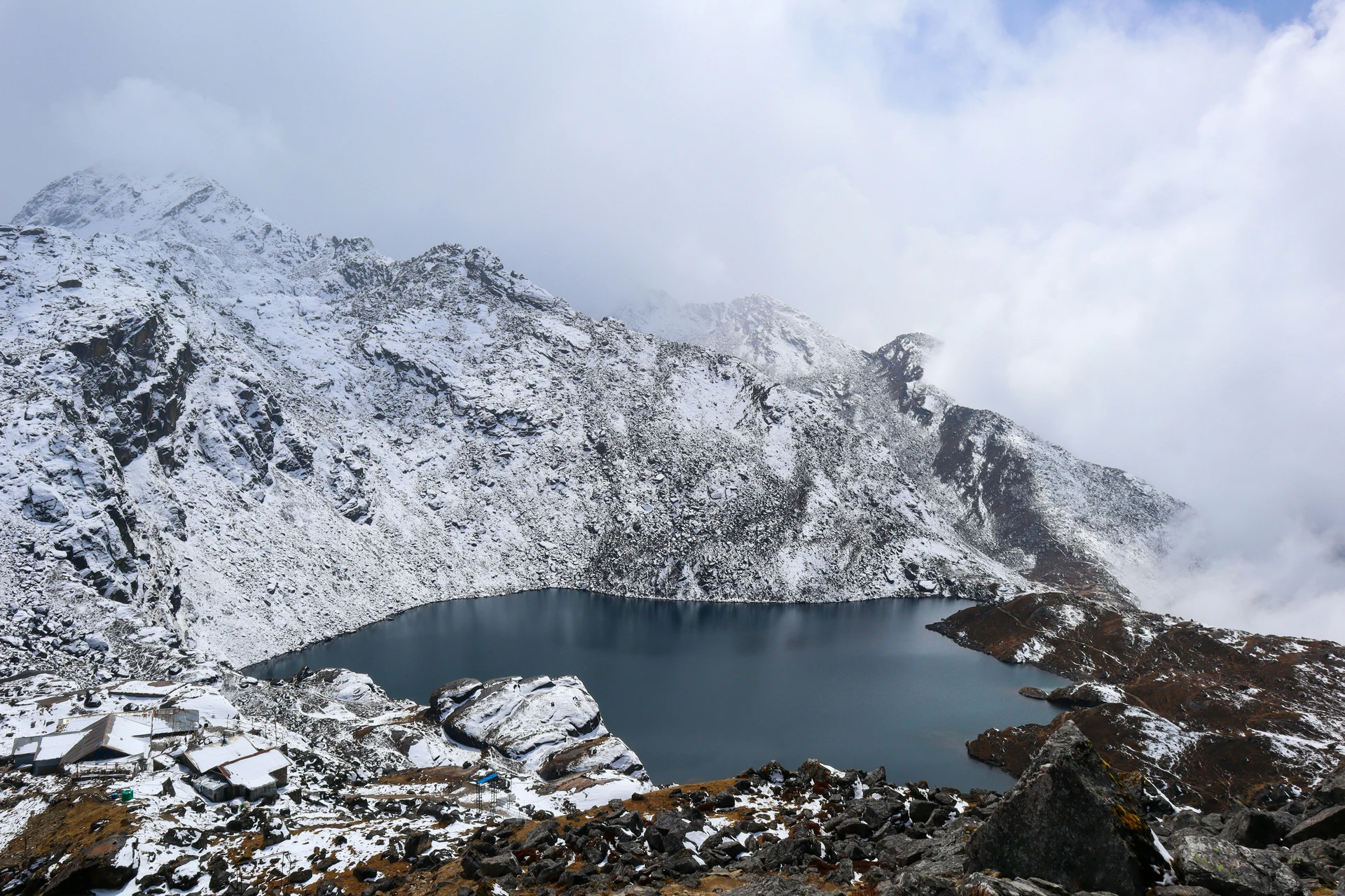 Gosaikunda Lake surrounded by snow covered mountains seen during cloudy day.