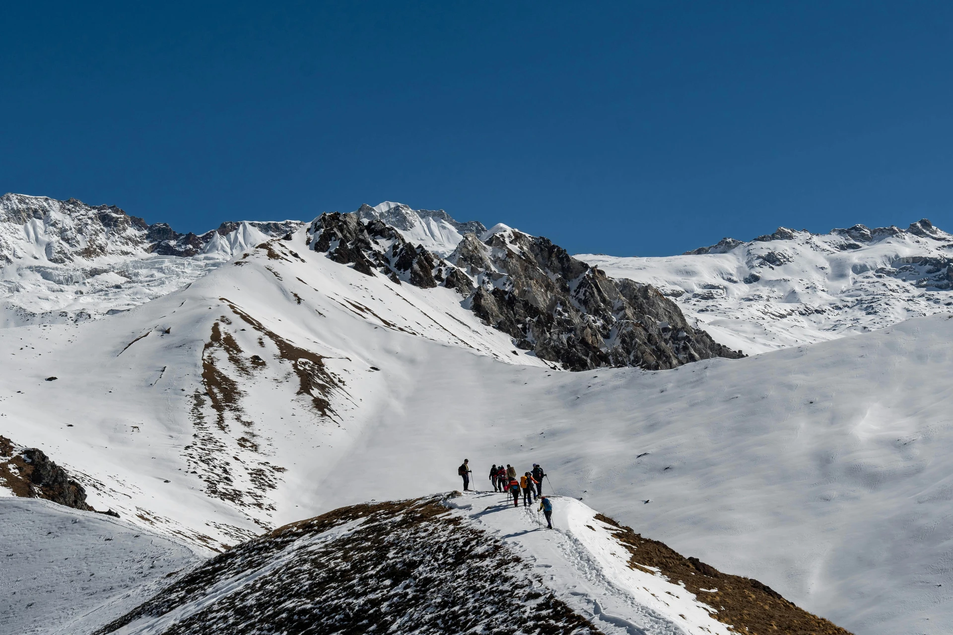 The image showcases a breathtaking mountainous landscape with a group of trekkers hiking along a snow-covered ridge seen in Langtang Gosaikunda Trek. The background consists of towering, snow-capped peaks under a clear blue sky. The terrain is a mix of snow and exposed rocky patches, indicating a high-altitude environment, possibly in the Himalayas or another major mountain range.