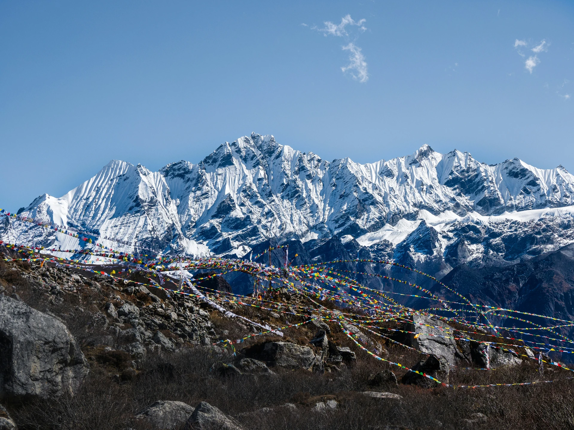This image captures a stunning mountainous landscape with snow-capped peaks in the background under a clear blue sky. In the foreground, colorful Tibetan prayer flags are strung across rocky terrain, adding a spiritual and cultural touch to the scene. The combination of rugged rocks, dry shrubs, and fluttering flags against the majestic mountains creates a peaceful yet powerful atmosphere, from Langtang Valley Trek.