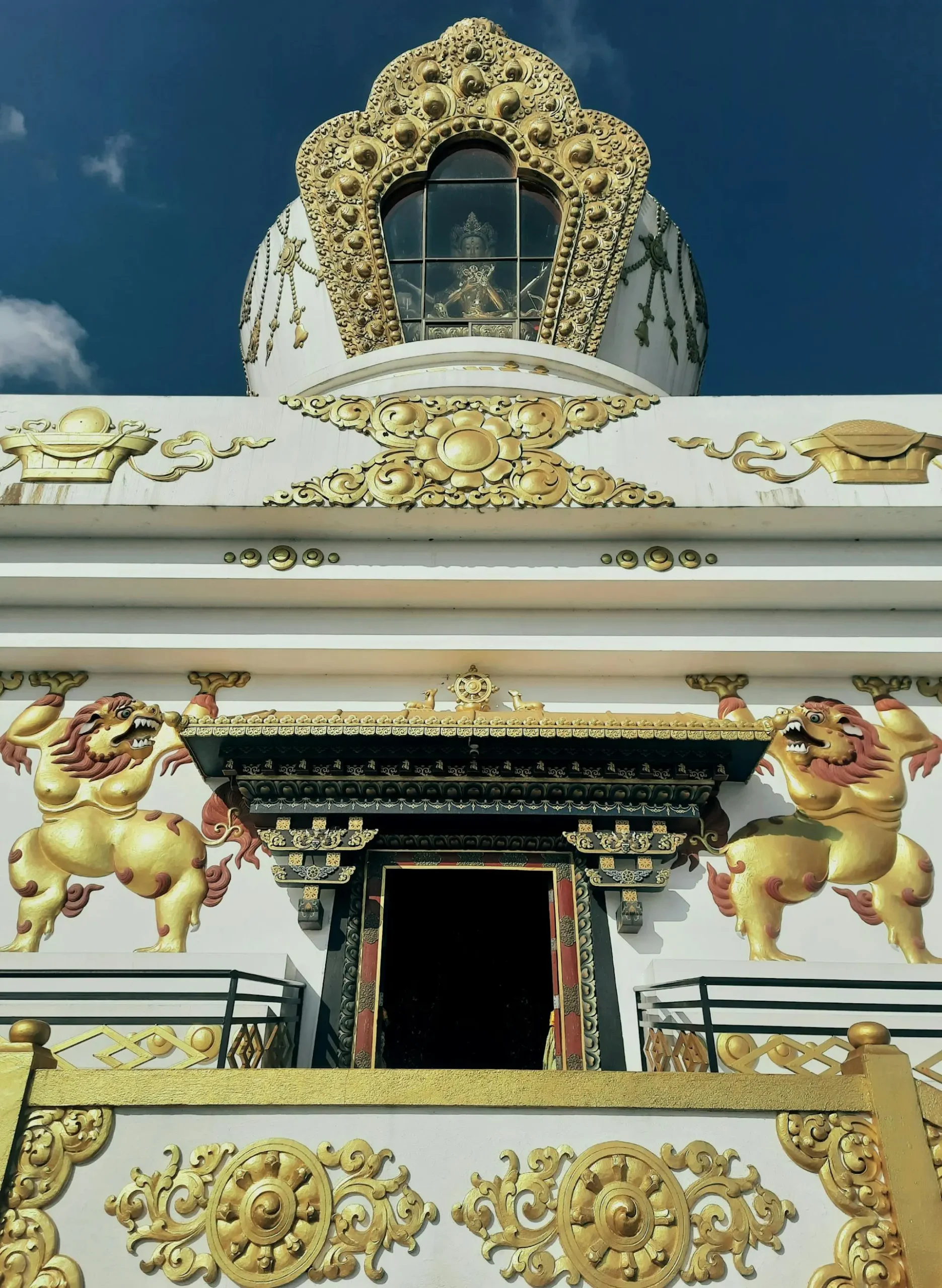 A beautifully decorated temple with intricate golden carvings and white walls, located on the Shivapuri-Budanilkantha route. The entrance is adorned with golden mythical lion figures, while a glass-enclosed deity statue sits at the top, framed by an ornate golden arch. The background features a clear blue sky.