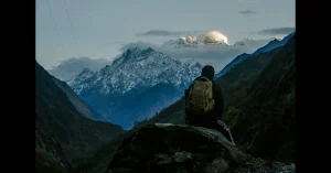 A traveler wearing a backpack sits on a rocky ledge, gazing at a breathtaking view of snow-capped mountains in Nepal. The golden sunlight reflects on the peaks, creating a serene and adventurous atmosphere. Which trekking route in Nepal are less crowded?