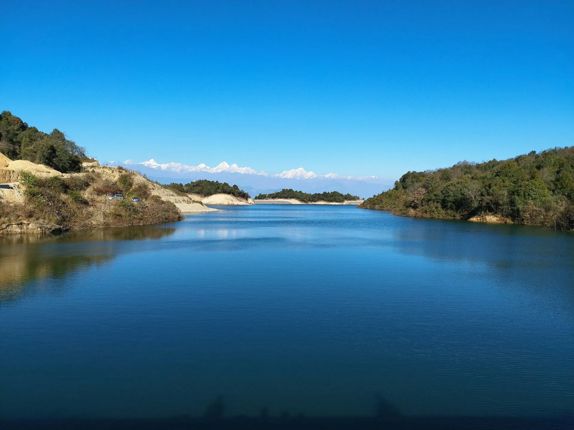 A scenic view of Dhap Dam in Shivapuri, Nepal, with its calm blue waters in the foreground. Lush green forests line the sides of the dam. In the background, a stunning mountain range of Nepal rises above tall hills. At the far end of the dam, another dense forest is visible. The sky is clear and bright, with no clouds, creating a beautiful and serene atmosphere.