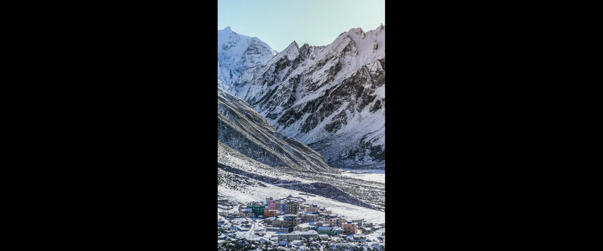 A picturesque view of Langtang Valley covered in snow, with colorful houses of Kyanjin Gompa nestled at the base of towering, rugged, snow-capped mountains under a clear sky.