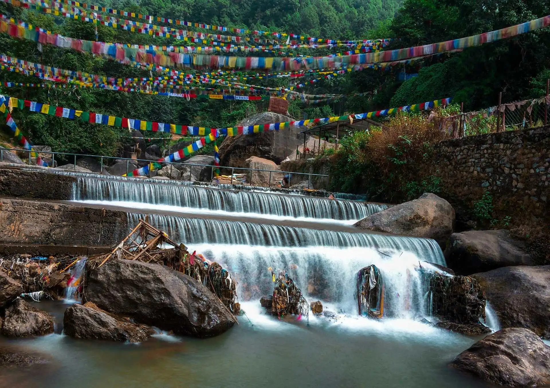 A serene cascading river in Shivapuri, Nepal, surrounded by lush green hills. Colorful Buddhist prayer flags are strung across the scene, adding a spiritual touch. Large boulders and traditional structures are visible near the water, while a small footbridge crosses the river in the background.