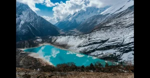 A breathtaking view from the Manaslu Circuit Trek in Nepal, featuring a vibrant turquoise glacial lake nestled among snow-draped mountains, with dramatic Himalayan peaks and rugged terrain under a sky filled with dynamic clouds—showcasing the remote beauty of this lesser-known trekking route.