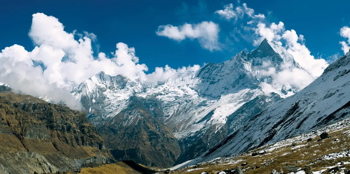 Snow-capped peaks of the Annapurna range pierce through dramatic clouds under a vivid blue sky, with rocky slopes and alpine terrain unfolding below.
