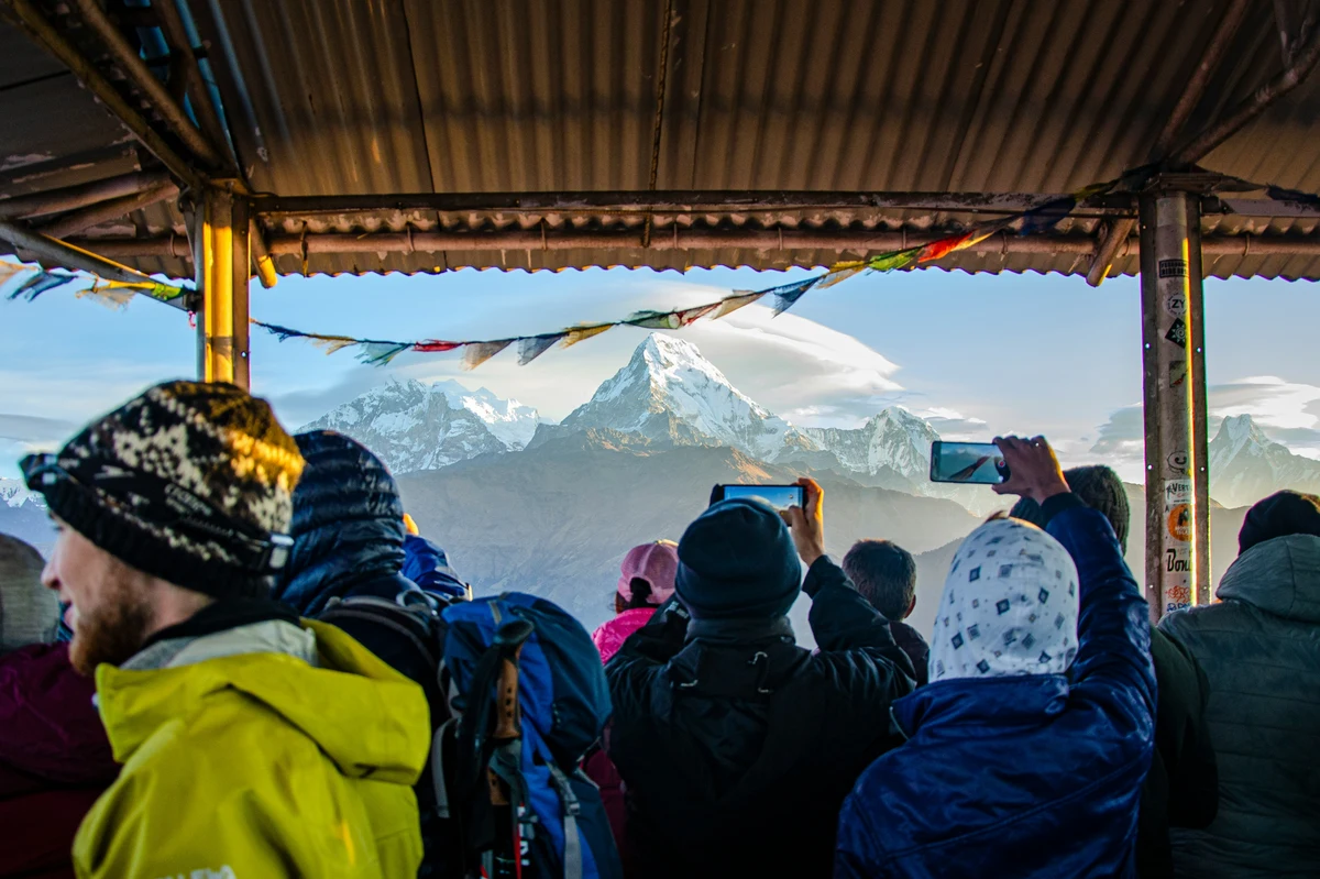 "Trekkers gathered under a shelter at Poon Hill, Nepal, capturing a stunning sunrise view of snow-capped Himalayan peaks with prayer flags fluttering overhead."