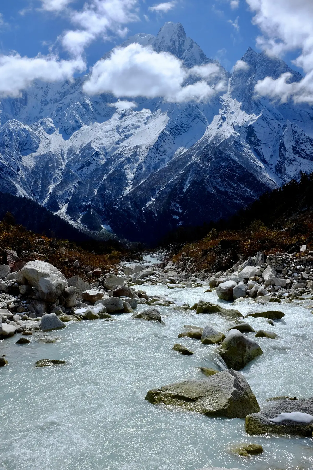 "Glacial river flowing over boulders along the Manaslu Circuit Trek, with snow-covered peaks and dramatic mountain ridges towering in the background under a partly cloudy sky in Nepal."