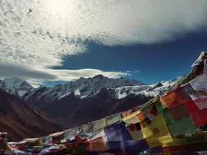 Colorful Tibetan prayer flags fluttering in the foreground with snow-capped mountains of the Langtang region in the background, under a dramatic sky — a serene moment capturing the essence of cultural trekking in Nepal.