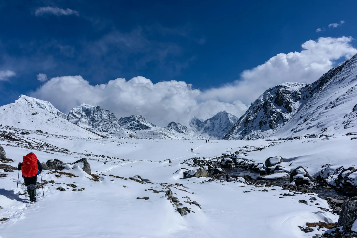 "Trekking in Nepal through a stunning snow-covered mountain landscape, with a trekker carrying a red backpack walking along a snowy trail surrounded by dramatic Himalayan peaks and a deep blue sky."