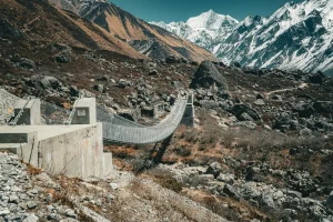  A sturdy suspension bridge stretches across a rocky Himalayan valley, flanked by rugged terrain and snow-capped mountains under a clear blue sky.
