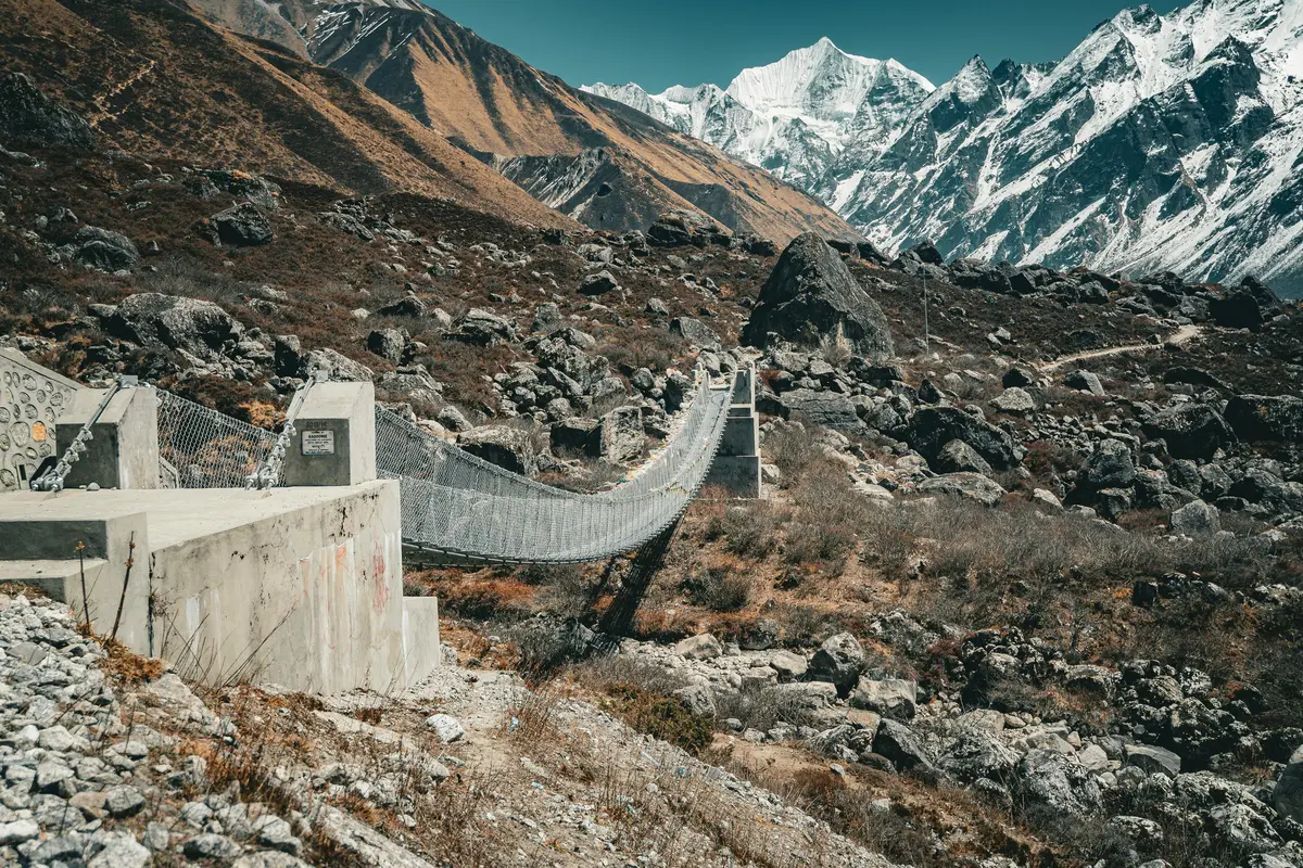 A sturdy suspension bridge stretches across a rocky Himalayan valley, flanked by rugged terrain and snow-capped mountains under a clear blue sky.