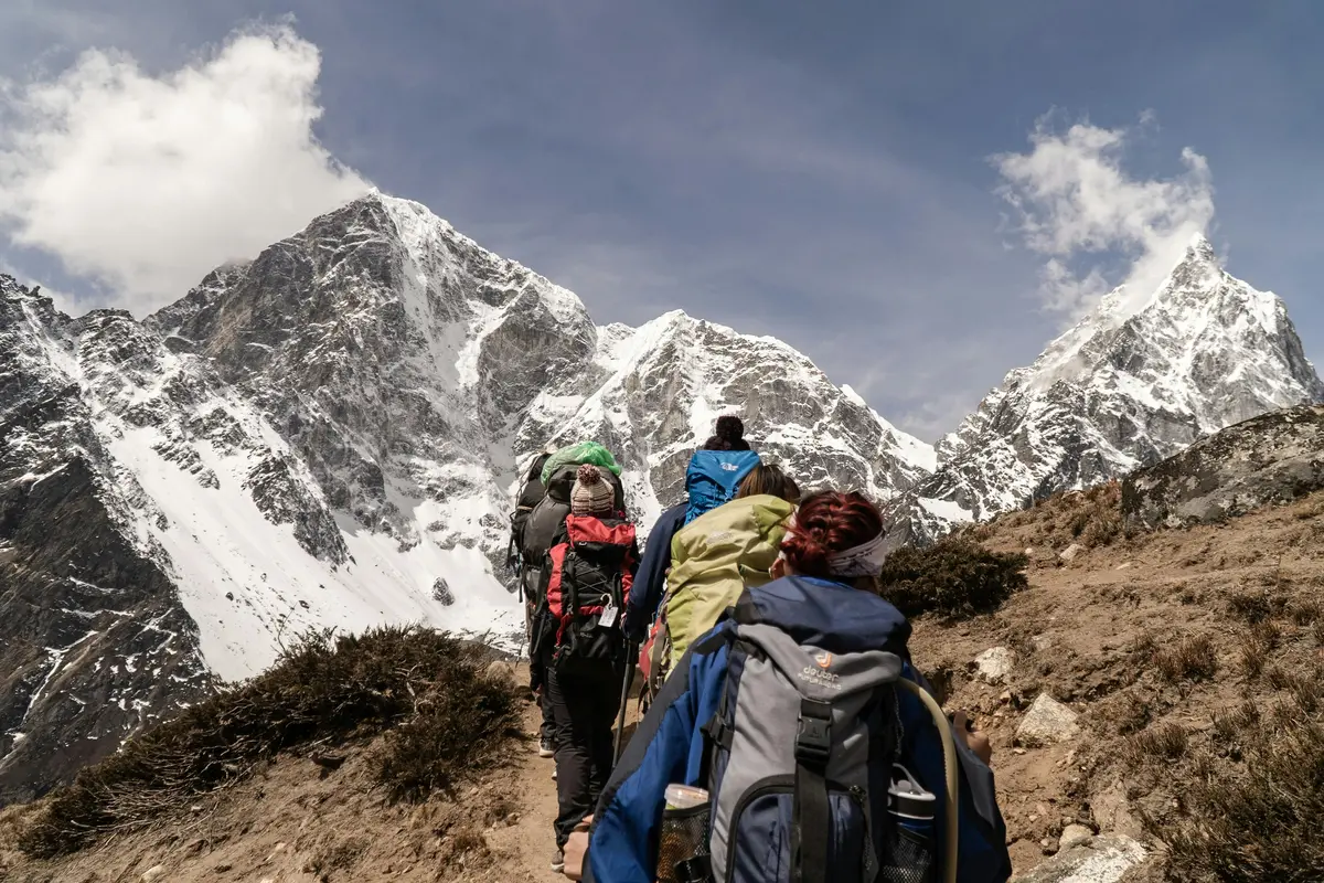 A group of trekkers ascend a rugged Himalayan trail with snow-covered peaks in the background, demonstrating travel responsibility by staying on designated paths and carrying their own gear to minimize environmental impact.