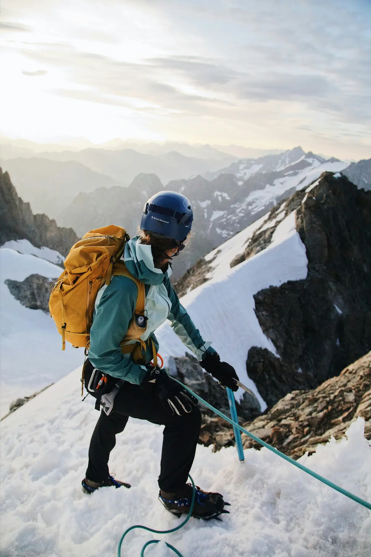 A mountaineer wearing a helmet, gloves, and a yellow backpack climbs a snow-covered ridge using an ice axe and rope, with rugged snow-capped peaks in the background during sunrise or sunset.