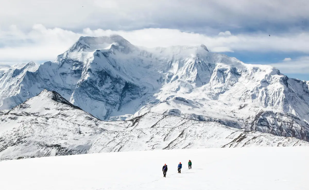 Four trekkers walk across a vast snow-covered landscape toward towering, majestic snow-capped mountains under a partly cloudy sky.