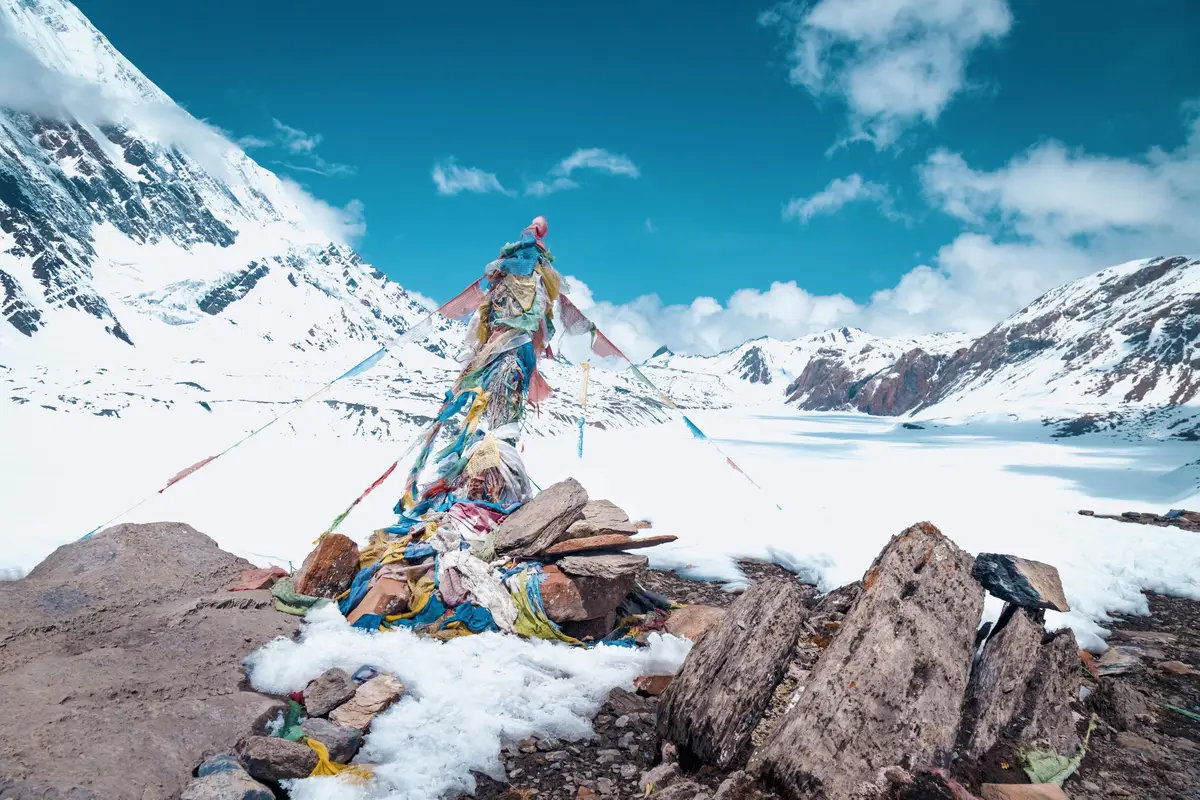 A high-altitude snow-covered mountain landscape featuring a pile of rocks adorned with colorful Tibetan prayer flags in the foreground, with a frozen lake and snow-clad peaks stretching into the distance under a bright blue sky with scattered clouds.