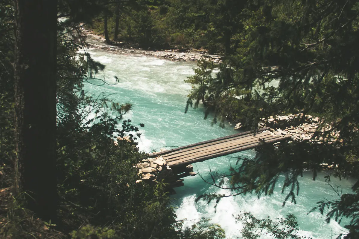"A wooden footbridge over a turquoise river surrounded by lush forest, symbolizing the serene and untouched beauty experienced while trekking in Nepal after COVID."