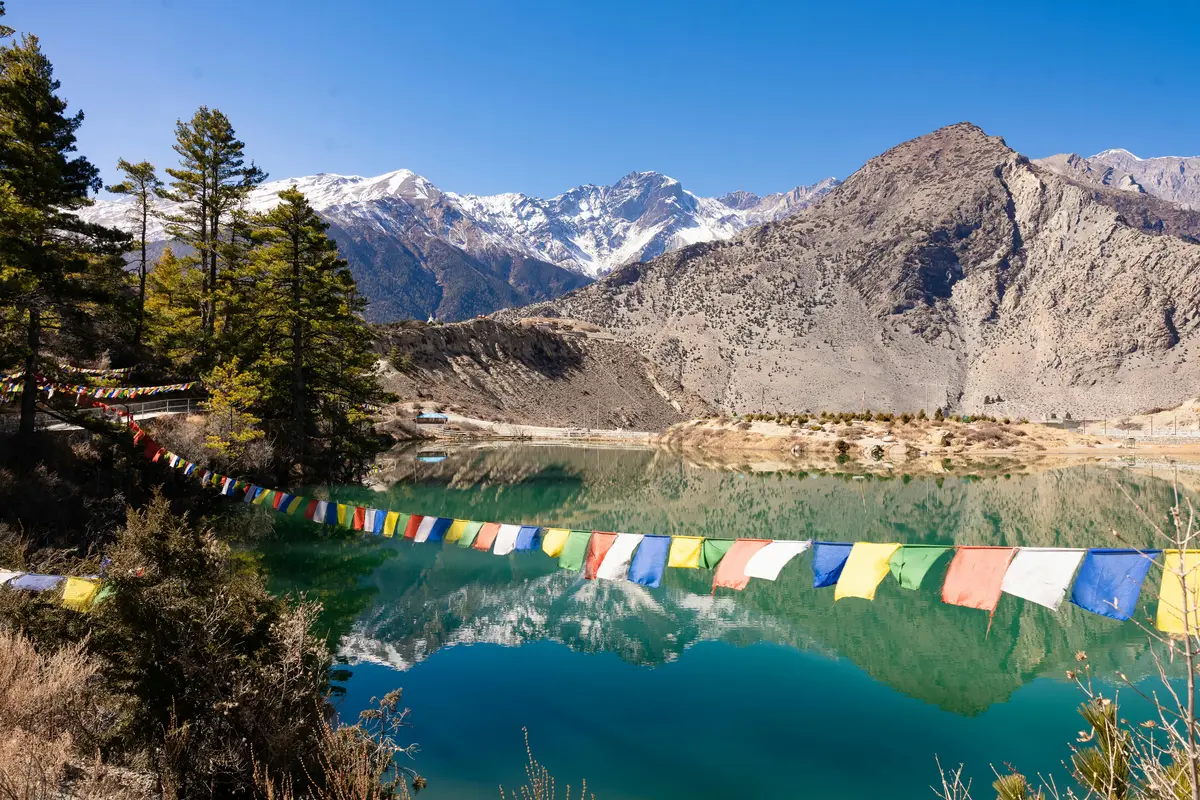 Colorful prayer flags flutter near the serene turquoise waters of Dhumba Lake, with snow-capped peaks and forested slopes reflecting in the lake, symbolizing the spiritual essence and natural beauty deeply rooted in Nepalese culture. 