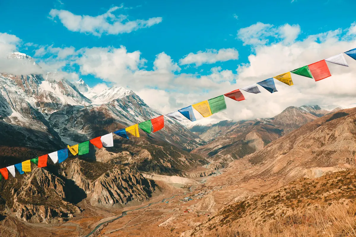 Colorful prayer flags fluttering in the wind against the backdrop of snow-capped Himalayan peaks and a remote mountain valley, symbolizing the deep spiritual connection and natural beauty rooted in Nepalese culture.