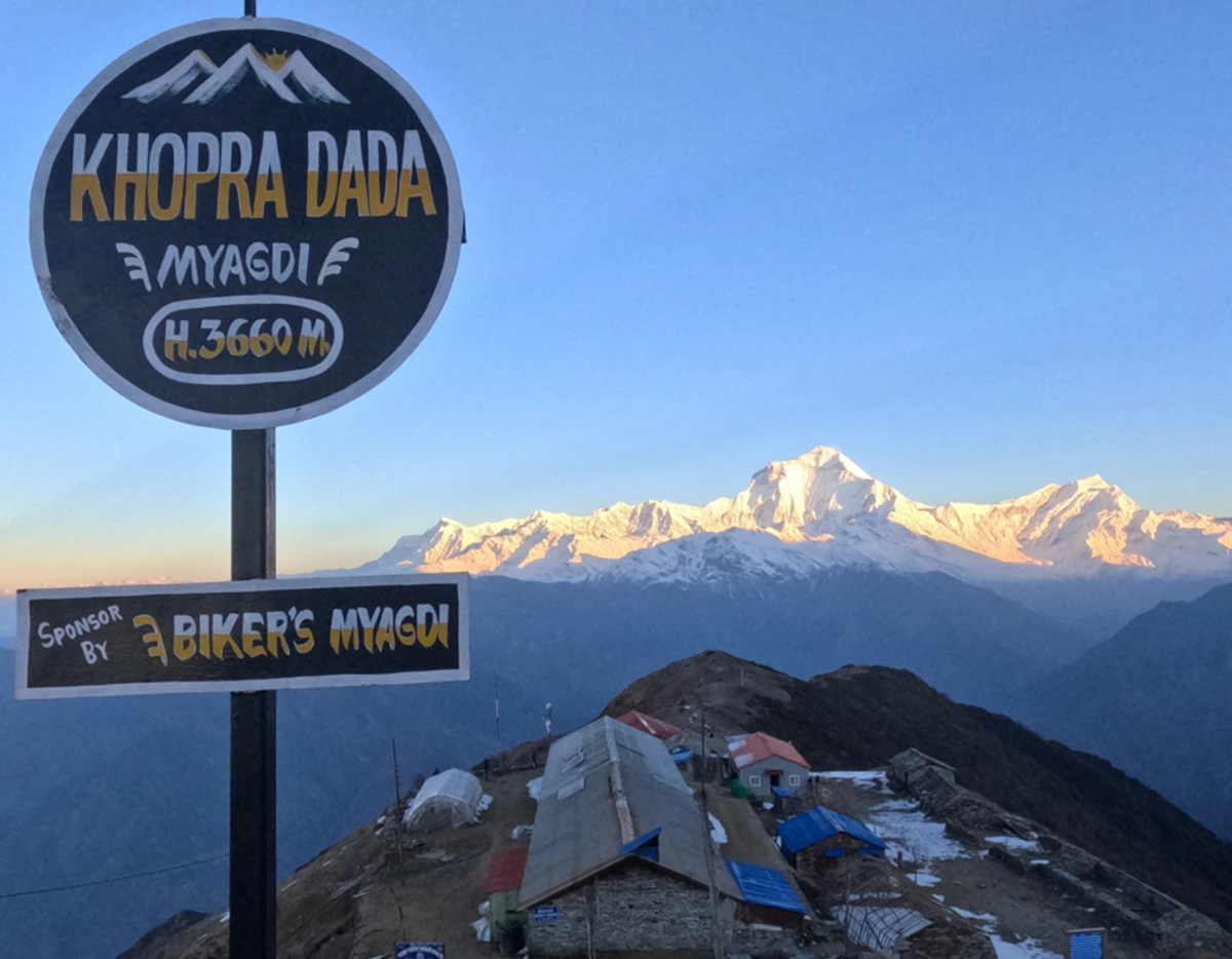 Trekker viewpoint at Khopra Danda (3,660m) with the local sign board and a clear view of Mount Dhaulagiri (8,167m) standing tall in the background seen during Khopra Ridge Trek Nepal.